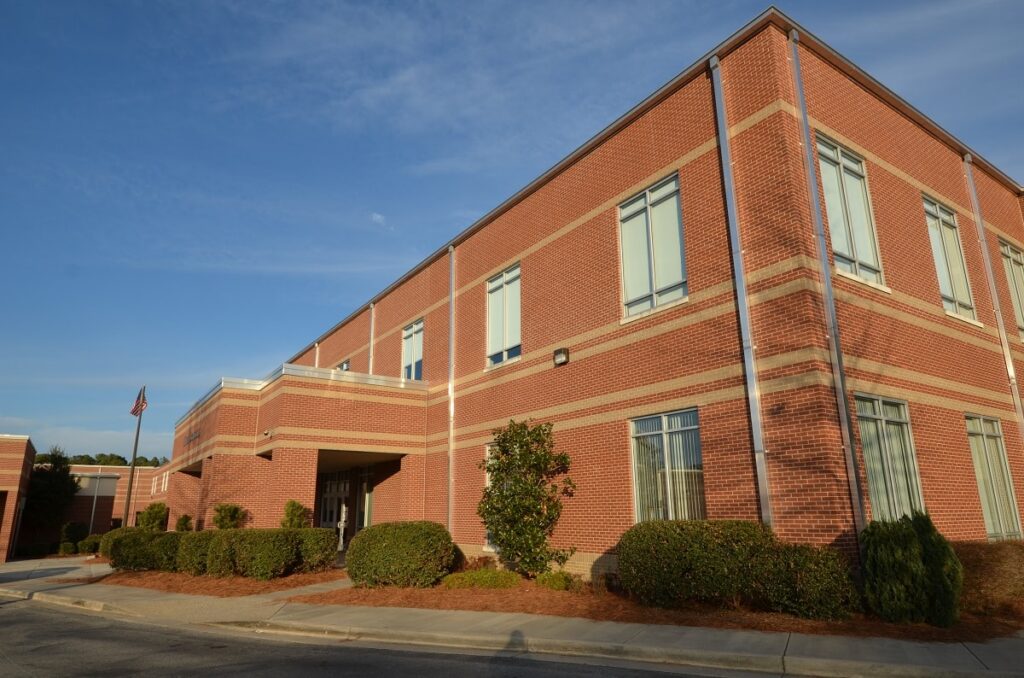 Two-story brick school building featuring numerous windows, bordered by neatly trimmed bushes and set beneath a clear blue sky.