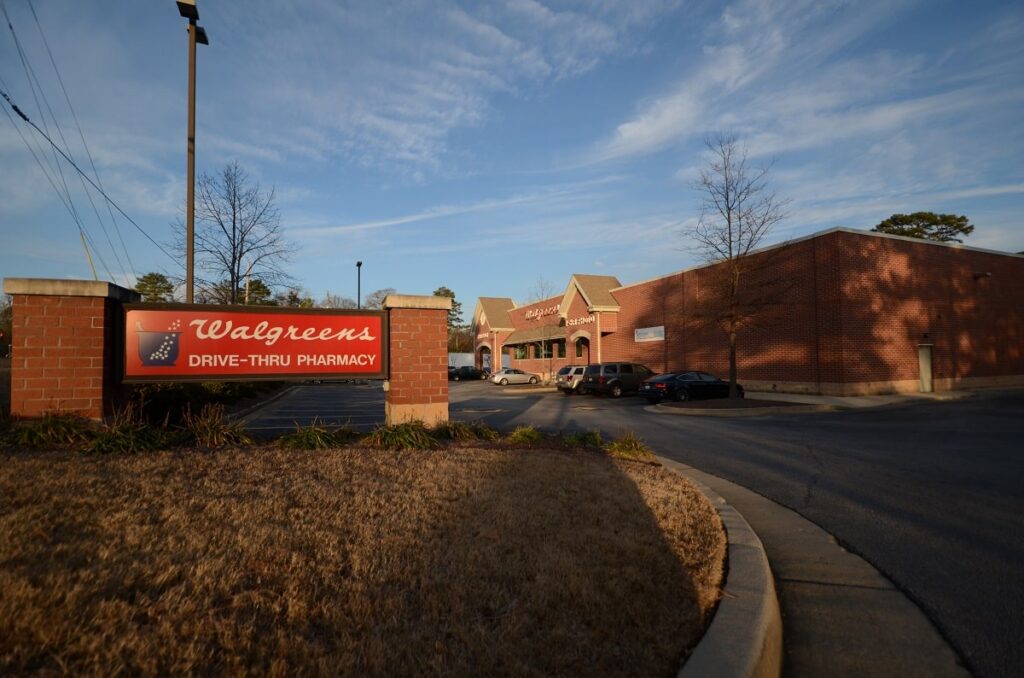 A Walgreens pharmacy building made of brick, featuring a drive-thru sign at the front, with a mostly empty parking lot and several leafless trees nearby beneath a partly cloudy sky.