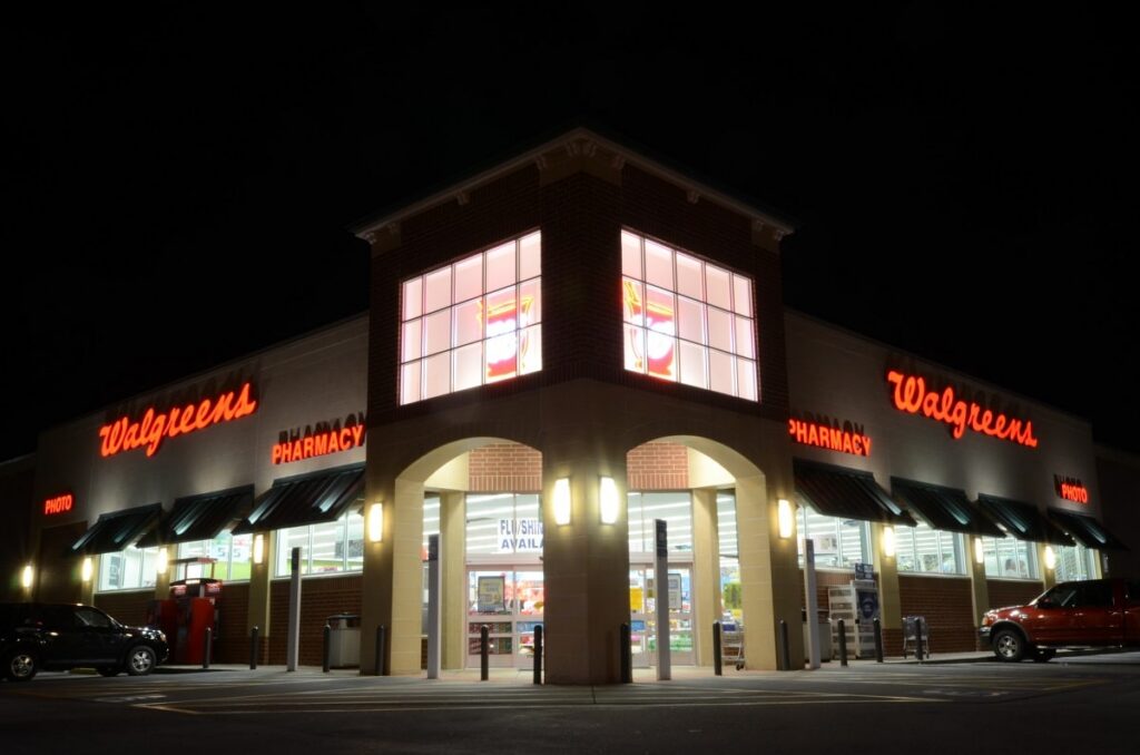 A Walgreens store at night featuring bright red neon signs, expansive windows, and a brightly lit entrance. Multiple cars are parked outside, and the glowing interior reveals the pharmacy and photo sections.