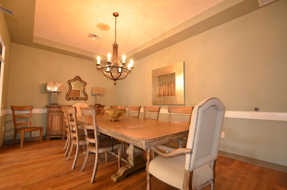 A dining room featuring a large wooden table surrounded by ten chairs, a chandelier overhead, artwork and a mirror on the soft green walls, lamps placed on a sideboard, and gleaming hardwood floors.