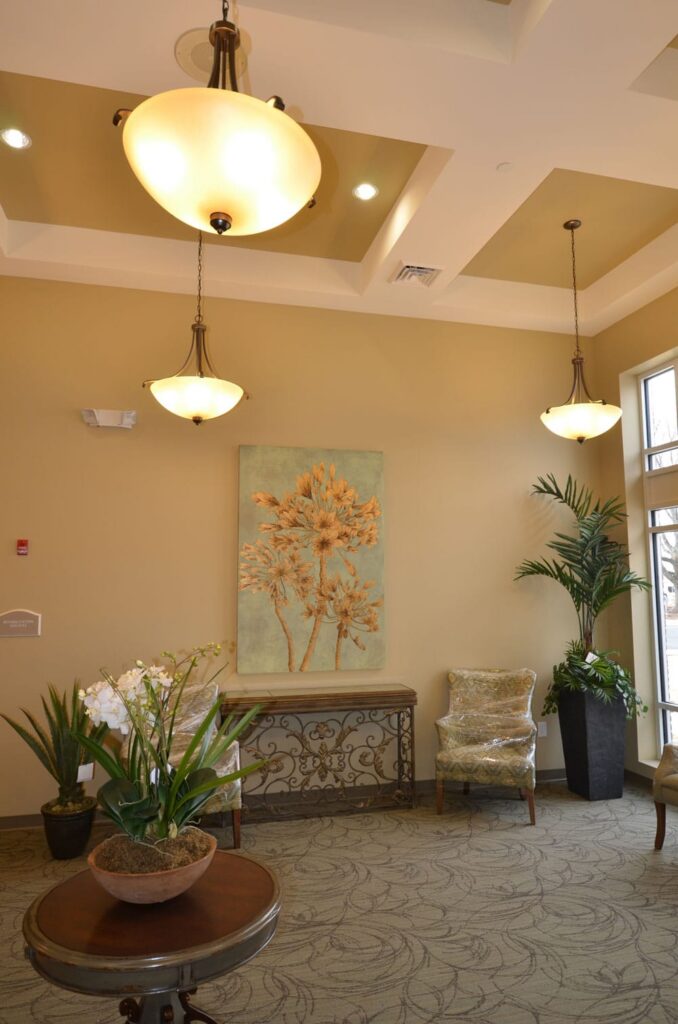 A warmly lit lobby featuring potted plants, a round table with two chairs, a decorative console table, floral artwork on the walls, and large windows that fill the space with natural light.