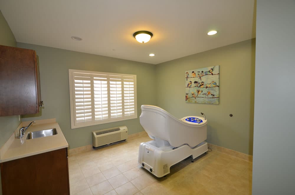 A spacious room features light green walls, a foot spa chair, a small sink, shuttered windows, an air conditioning unit, and a painting depicting swimmers on the wall.