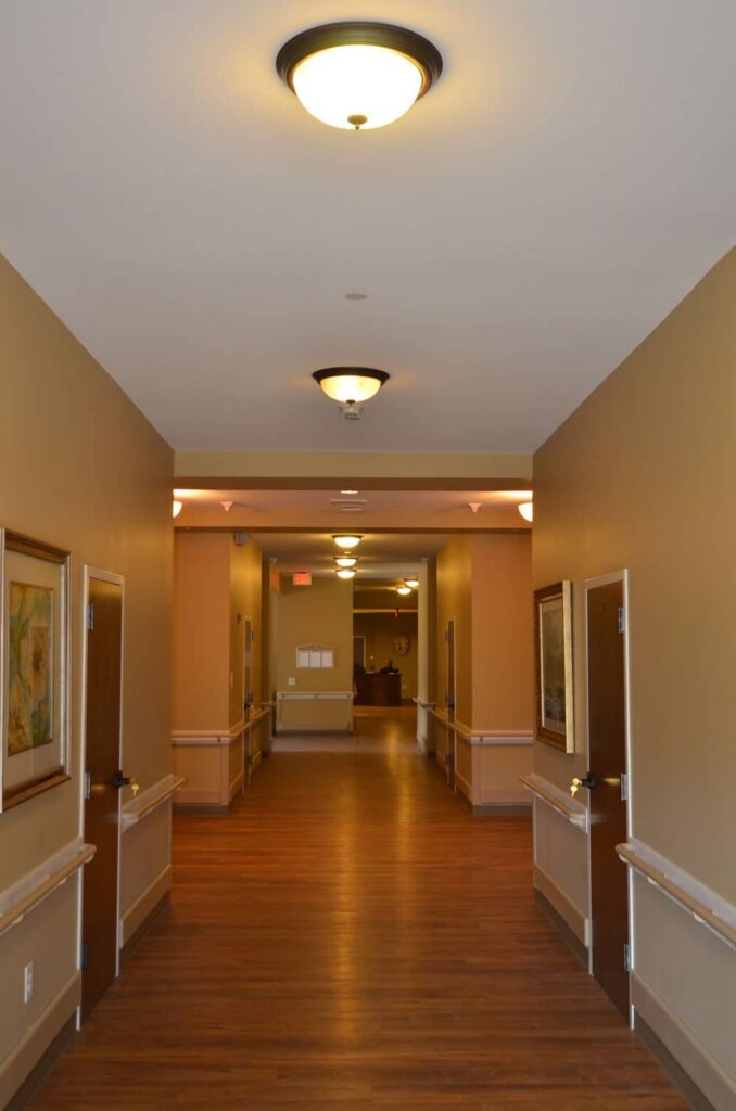 Bright hallway featuring wooden floors, tan-painted walls, sturdy handrails, several closed doors, and framed artwork hung along both sides, extending toward an open area at the far end.