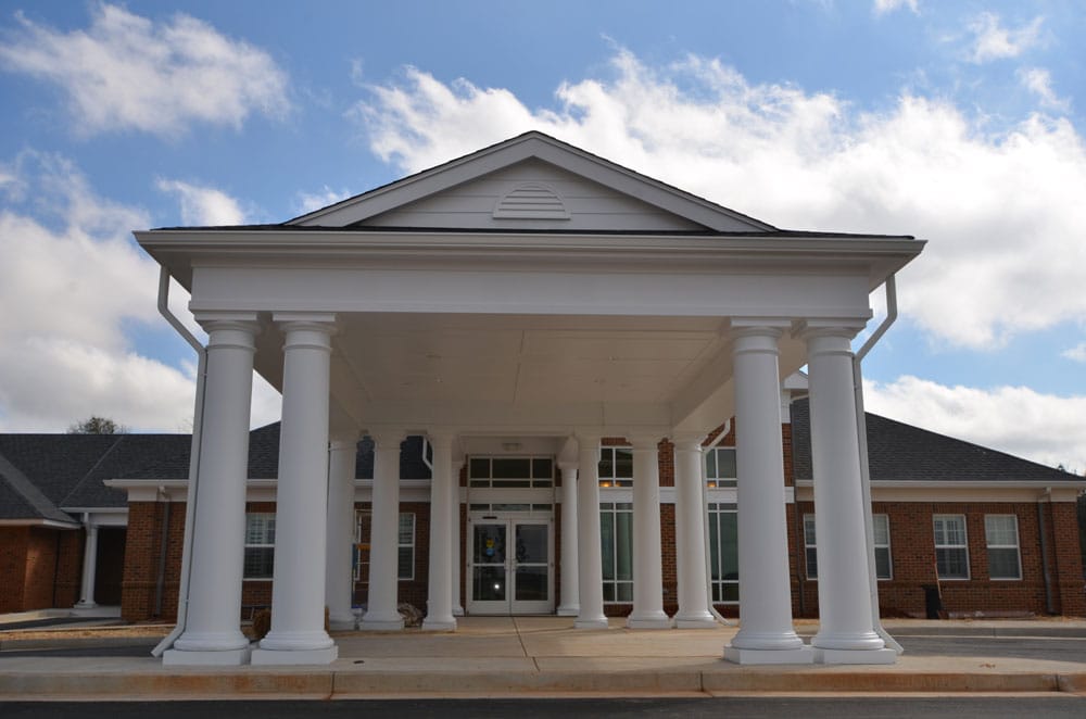 The entrance of a building featuring tall white columns that hold up a covered portico, with expansive windows and brick walls beneath a sky dotted with some clouds.