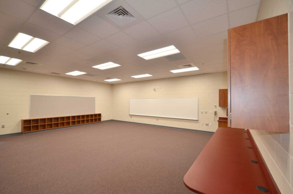 Empty classroom featuring a carpeted floor, two large whiteboards, cubby shelves, fluorescent ceiling lights, and a wooden counter along one wall. The walls are light-colored and the room contains no chairs or desks.