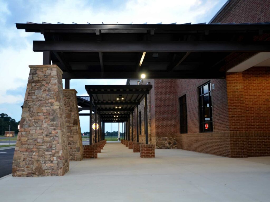 A covered walkway features stone pillars and brick walls, guiding visitors toward the entrance of a building that appears to be a school or public facility in the early evening.