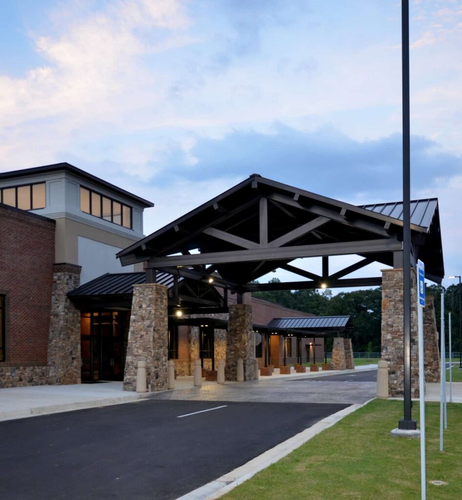 A contemporary building featuring a covered drive-through entrance upheld by stone columns, expansive windows, and a brick facade, all beneath a sky with scattered clouds.