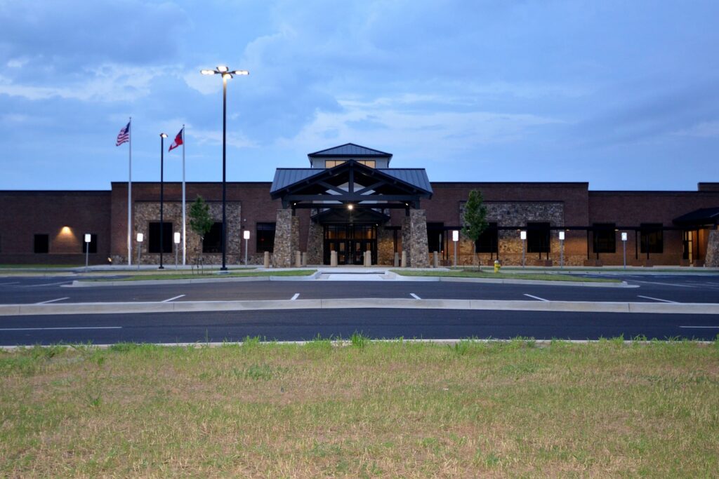 A modern single-story brick building featuring a large entrance and three flagpoles stands in front of an empty parking lot beneath a cloudy evening sky, with grass visible in the foreground.