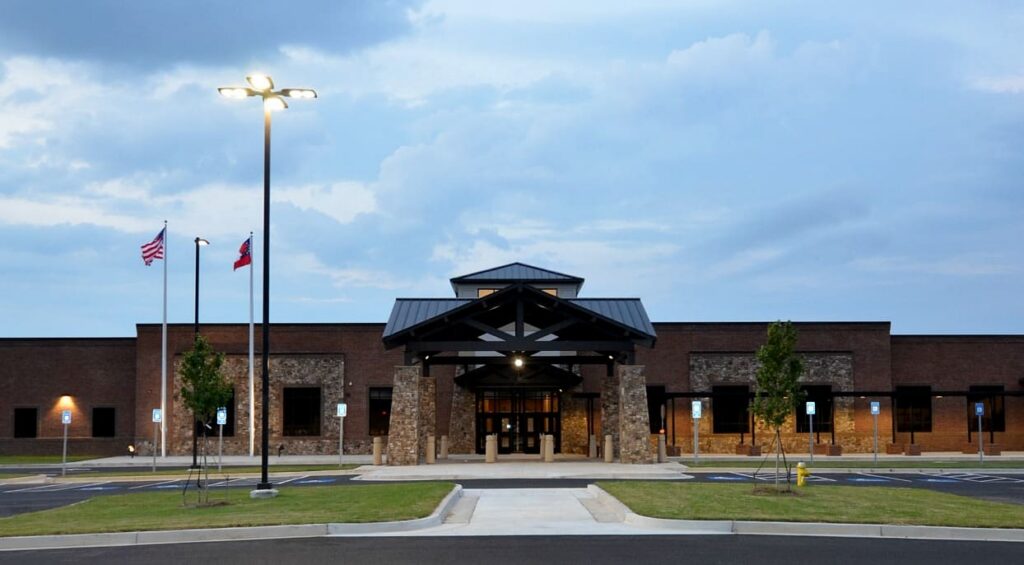 A modern brick building with a covered entrance, two flagpoles flying the U.S. and state flags, and an empty parking lot beneath a cloudy dusk sky.