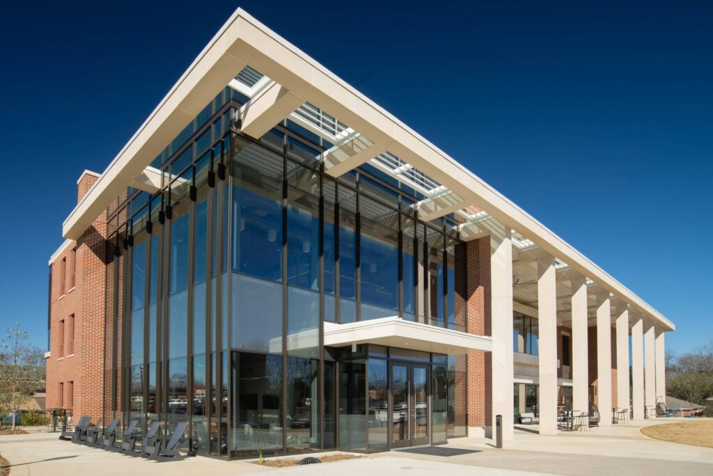 Modern building featuring expansive glass windows, brick walls, and tall white columns beneath a clear blue sky. Outdoor patio seating is arranged near the entrance.