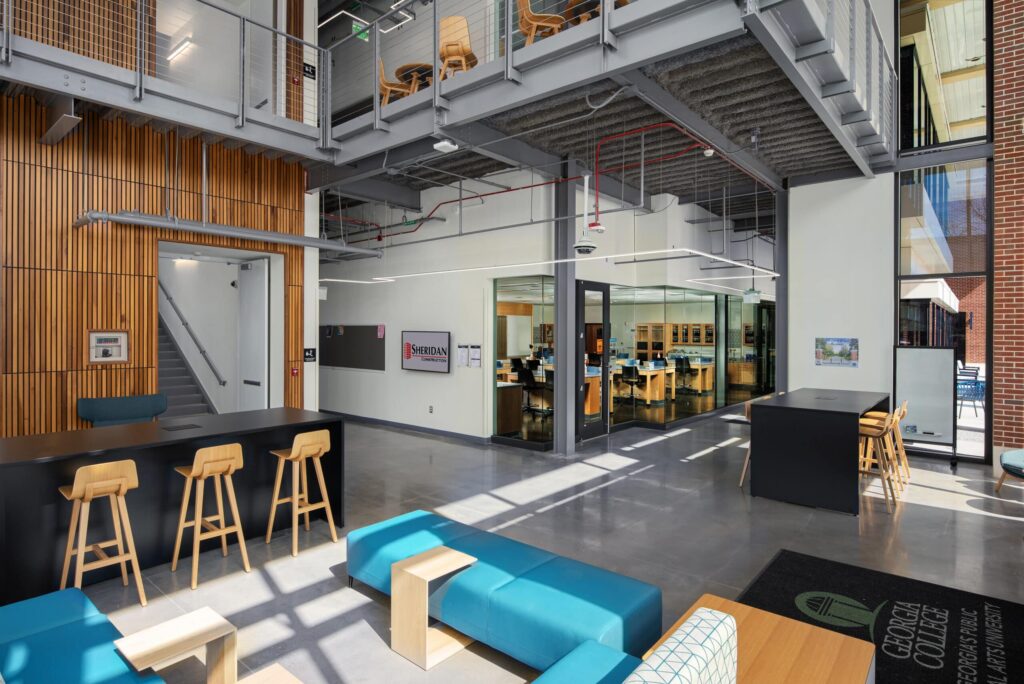 A contemporary lobby features teal sofas, wooden stools, and high tables, along with a glass-walled room and wood paneling. There’s an upper walkway, and sunlight pours in through expansive windows, creating an open and welcoming atmosphere.