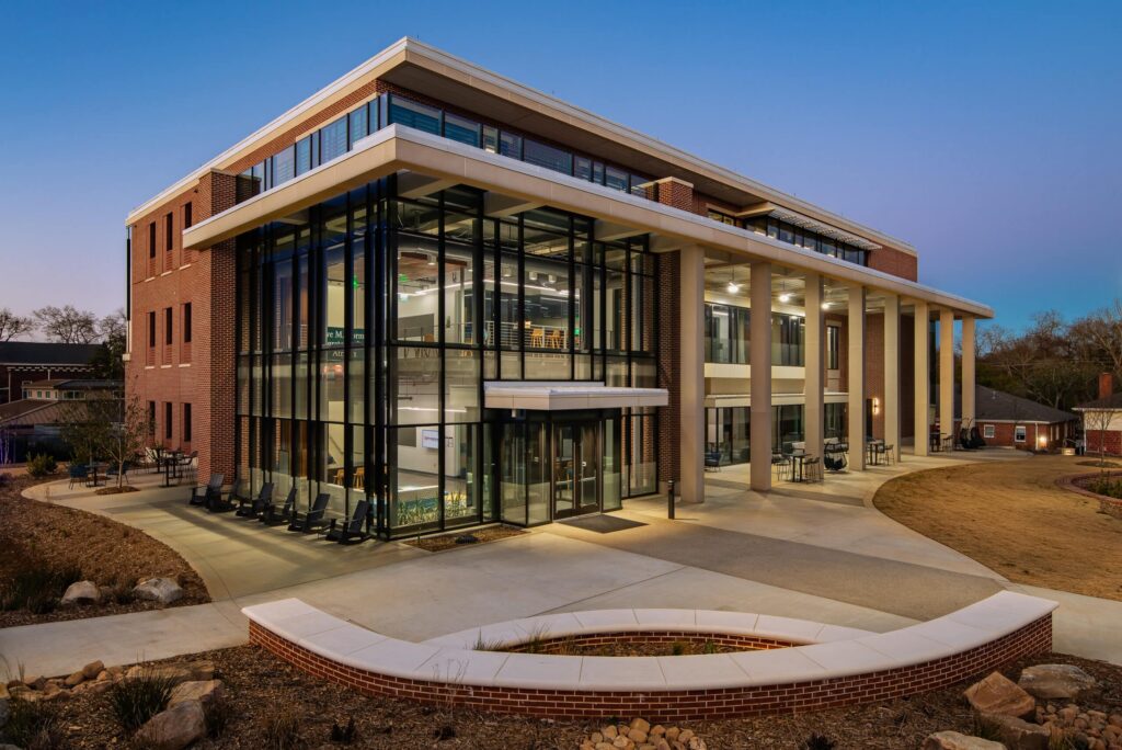 Modern three-story building featuring expansive glass windows, a brick exterior, and tall columns, all illuminated during dusk. The structure is surrounded by landscaped pathways.