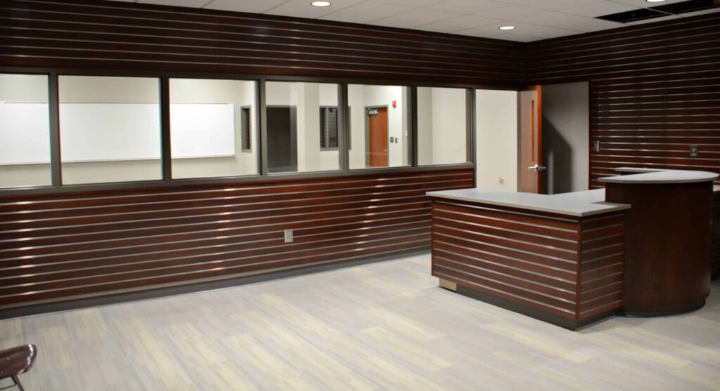 Reception area featuring dark wood paneling, a curved desk, and large glass windows that reveal an adjacent room with a whiteboard. The space has light wood flooring and is illuminated by fluorescent lights.