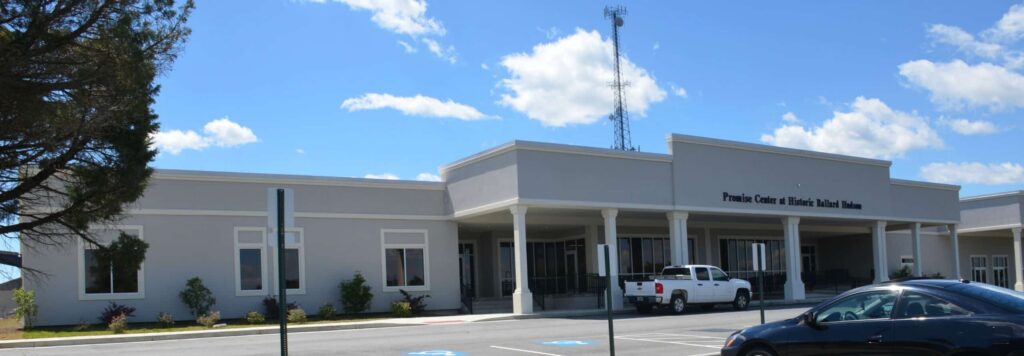 A single-story building with columns at the entrance, labeled Promise Center. A white truck and a black car are parked in front, with blue sky and clouds in the background.