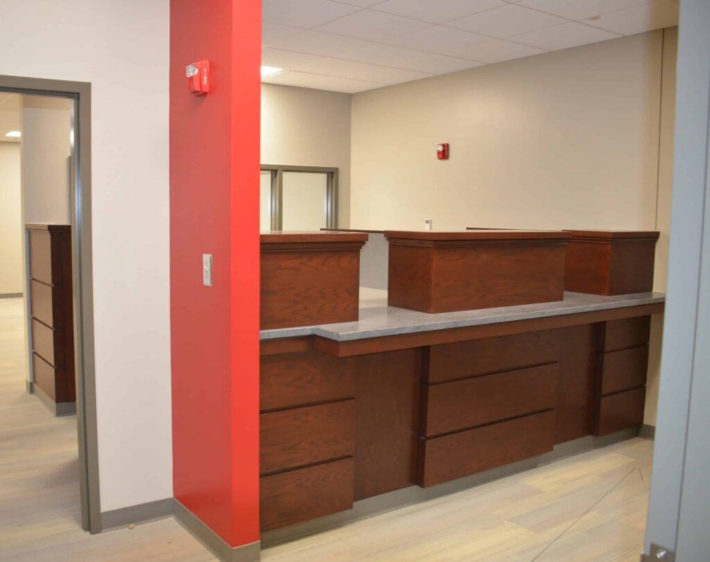 A modern office reception desk with a dark wood finish and a concrete countertop stands amid beige walls and a red accent wall. The space appears clean and is brightly lit by both natural and ceiling light.