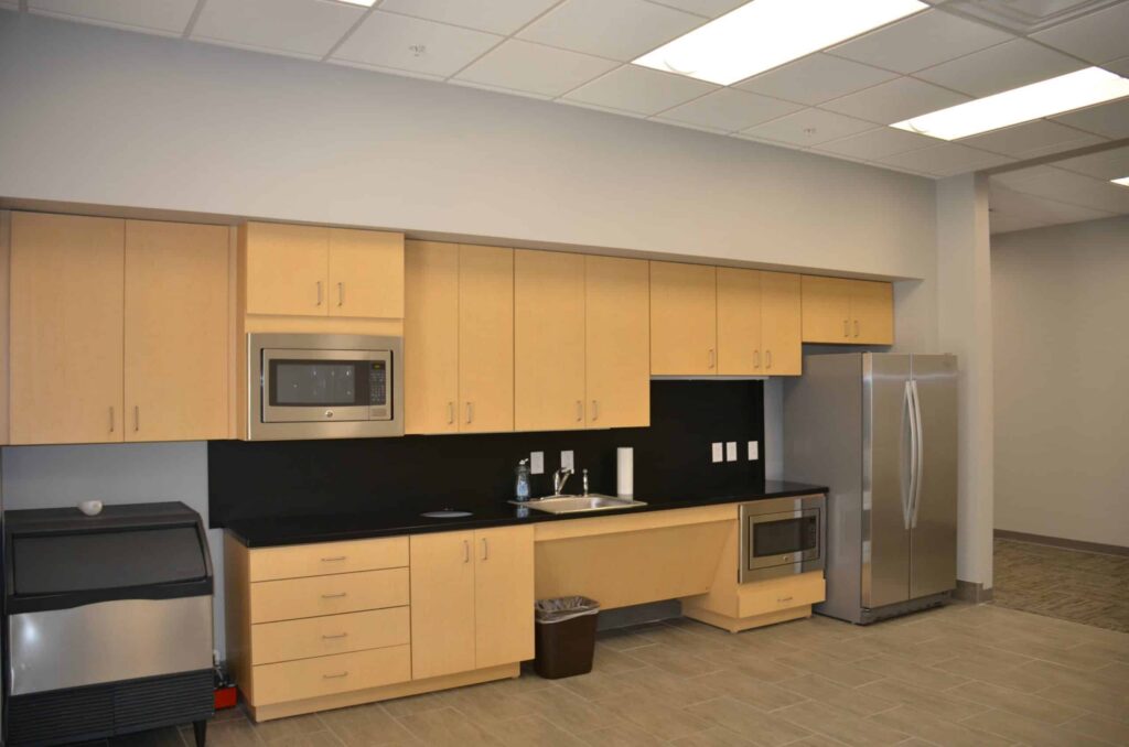 Modern office kitchen with light wood cabinets, a microwave, sink, trash can, stainless steel refrigerator, and ice machine. The room has tile flooring and ceiling lights.