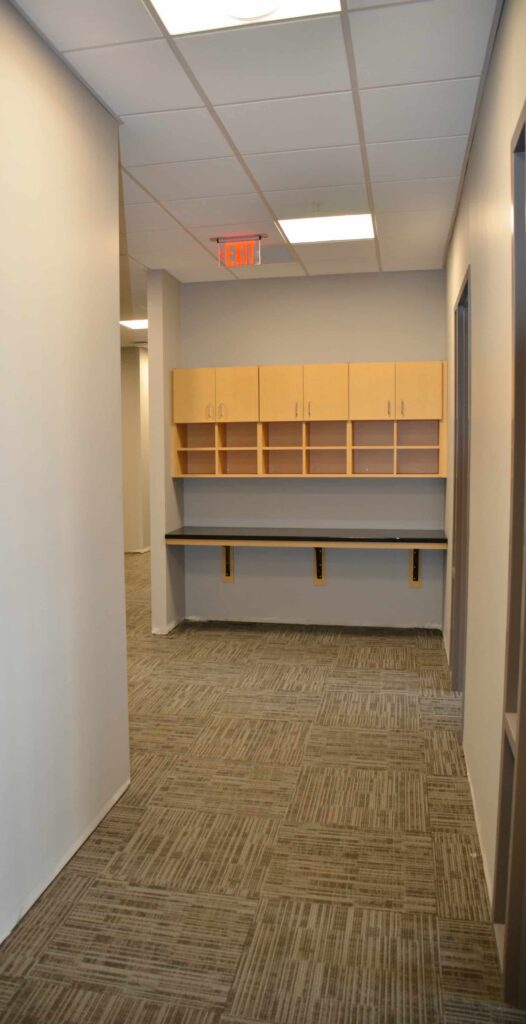 A hallway featuring gray carpet, beige walls, a black counter topped with wooden cabinets, and an exit sign mounted on the ceiling.