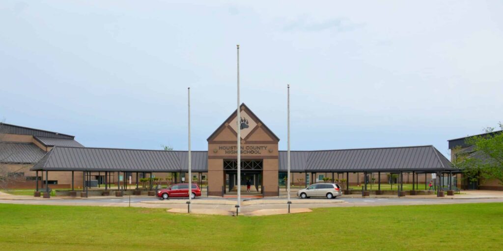 A front view of Houston County High School, featuring a covered walkway, two flagpoles without flags, and two cars—one red and one white—parked in front of the building on a cloudy day.