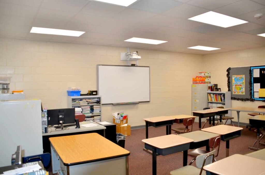 A classroom featuring empty desks, a whiteboard, and a teacher’s desk equipped with a computer. Bookshelves line the wall and various classroom supplies are present. The space is brightly lit by fluorescent lights and has beige-colored walls.