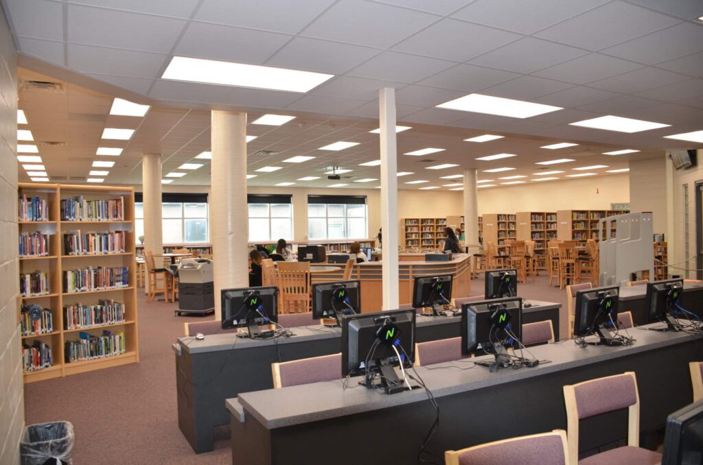 A spacious library features bookshelves, study tables, and multiple desktop computers on desks. Large windows fill the room with natural light, where a few people are reading or working. The ceiling displays rows of bright lights.