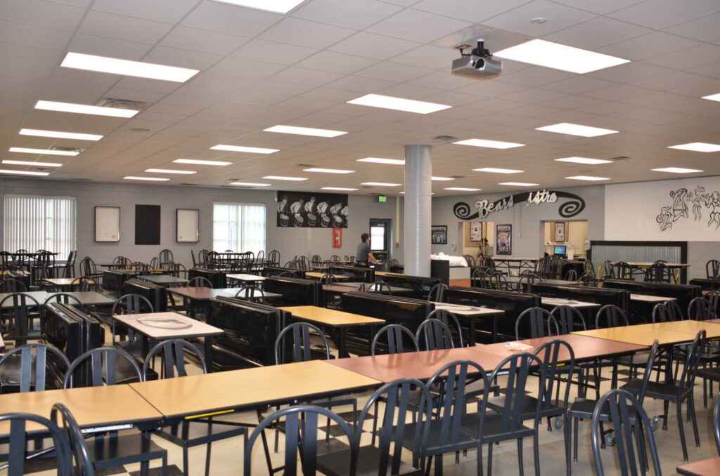 A spacious, empty cafeteria features rows of tables with black chairs beneath ceiling lights. Colorful artwork and various signs adorn the walls, while large windows allow natural light to brighten the space.