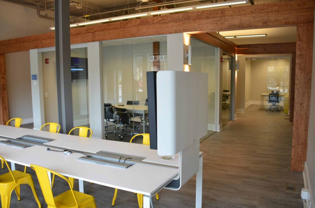 Modern office with a long white desk, yellow chairs, glass-walled meeting rooms, wood accents, and ceiling lights; monitors and computers are on the desk.
