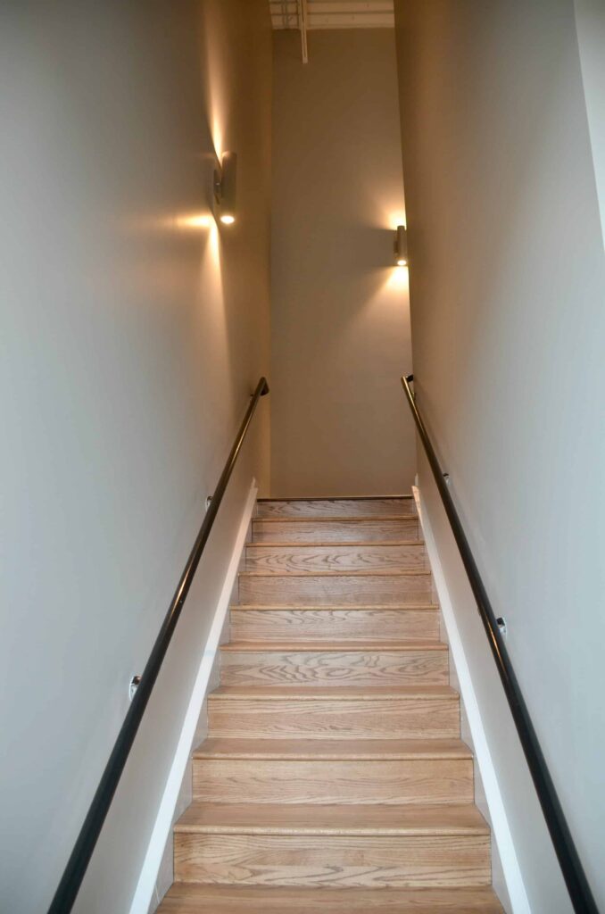 A wooden staircase with black handrails on both sides ascends between light gray walls, illuminated by modern wall-mounted lights.