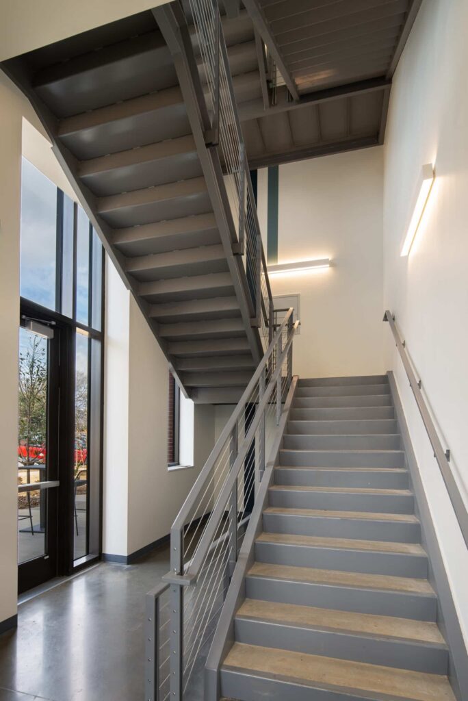 Gray metal staircase inside a building with natural light streaming through large windows on the left, a concrete floor, and sleek modern wall-mounted lights.