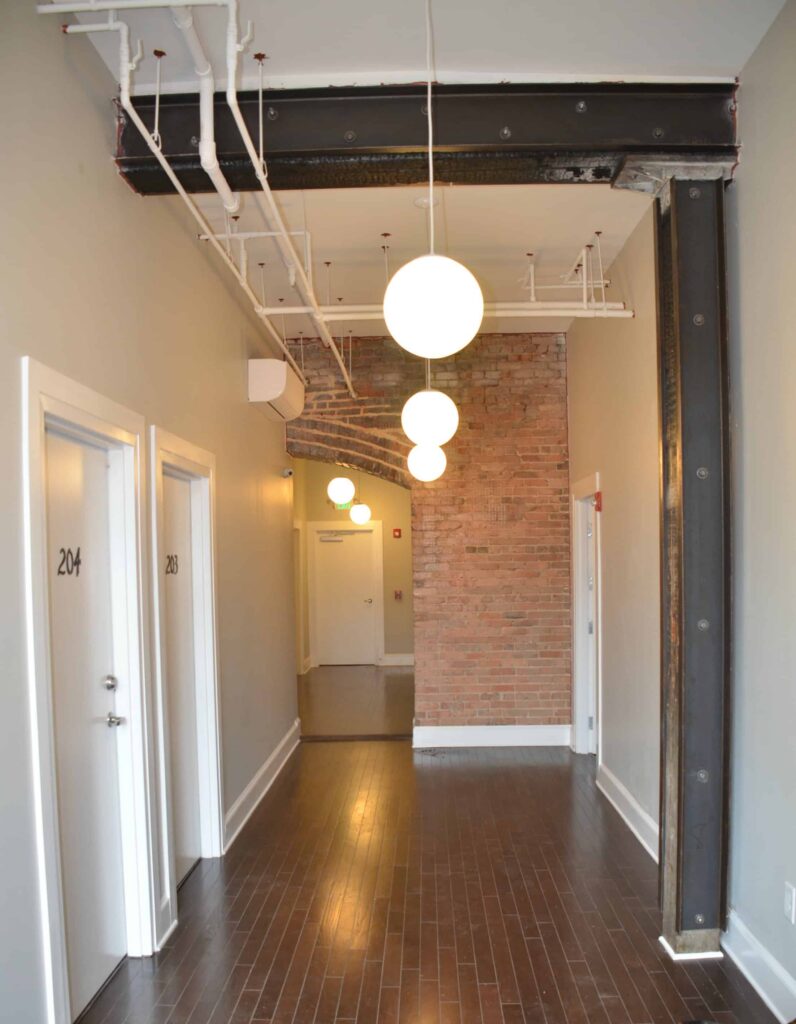 A hallway with wooden floors, white doors labeled 203 and 204, exposed brick wall, steel beam, and round ceiling lights hanging in a row.