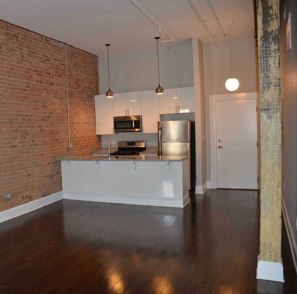 Modern kitchen featuring stainless steel appliances, white cabinets, a granite countertop, exposed brick wall, dark wood floors, and pendant lighting within an open-concept apartment.