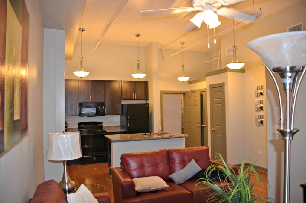 Modern apartment featuring an open floor plan. The cozy living room has red sofas, while the kitchen area includes dark cabinets and black appliances. Pendant lights hang from the ceiling, and a ceiling fan is overhead.