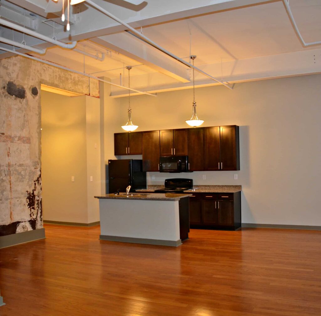 Modern kitchen featuring dark wood cabinets, black appliances, an island with a sink, pendant lights, hardwood floors, high ceilings, and an exposed concrete wall on the left. The space has a spacious, open layout.
