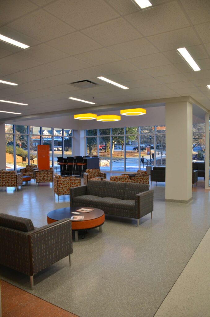 A modern lounge area featuring patterned sofas, round tables, large windows, and yellow ceiling lights. The spacious room is filled with natural light, and chairs are arranged to encourage socializing or relaxing.