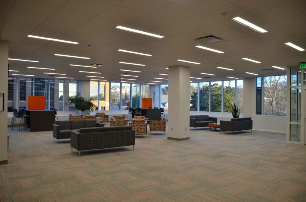 Spacious office lounge featuring modern sofas, armchairs, large windows allowing abundant natural light, with plants and partition panels adding decoration to the open, carpeted area.