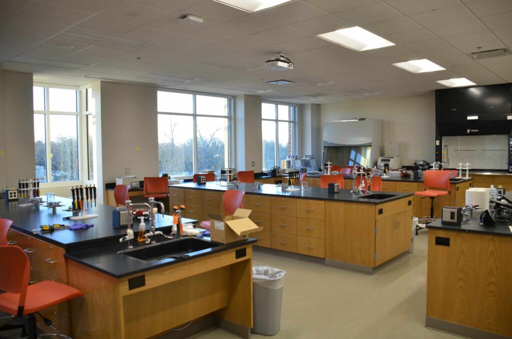 A modern science laboratory featuring wooden cabinets, black countertops, red chairs, sinks, pipettes, and large windows that allow natural light to fill the space.