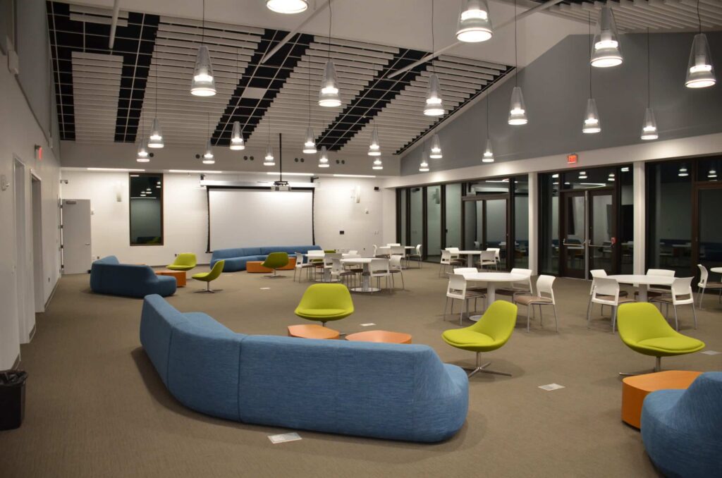 A modern lounge featuring blue sofas, green chairs, white tables, and pendant lights hanging from the ceiling. One wall has large windows and a projector screen. The space is open and neatly organized.