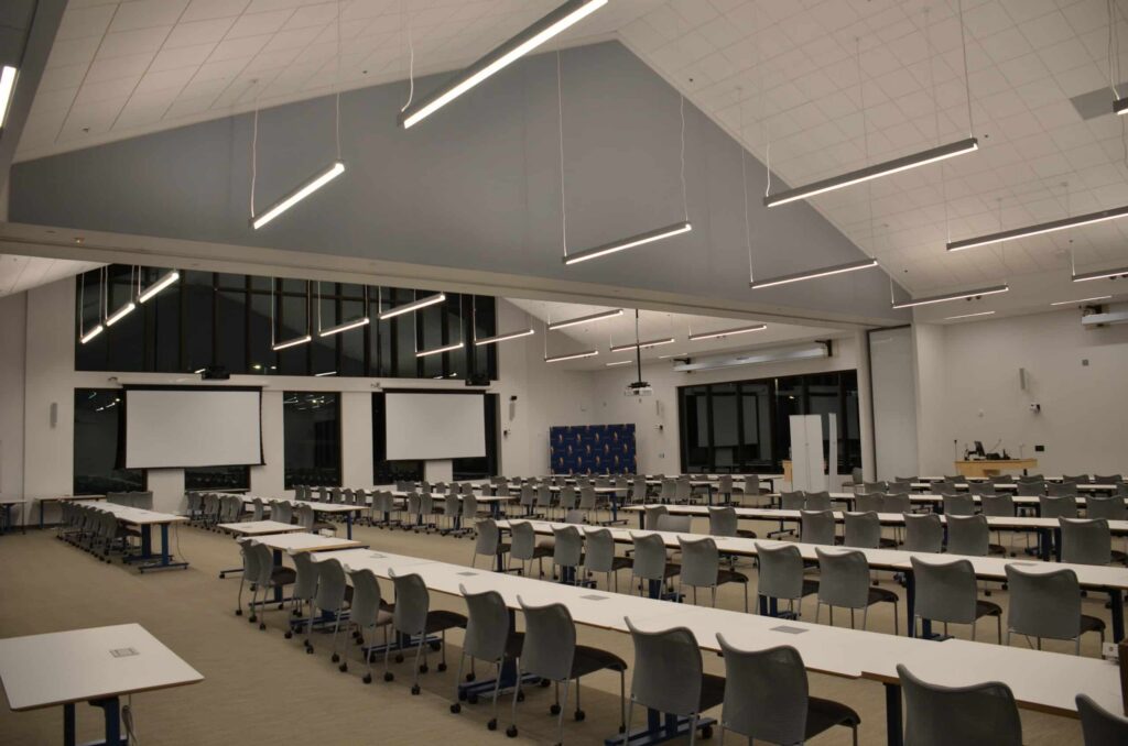 A spacious, contemporary conference room featuring rows of tables and chairs, two projector screens at the front, bright overhead lighting, and expansive windows along the rear wall. The room is empty and filled with natural light.