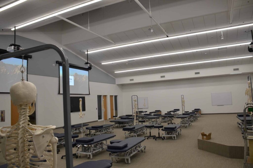 A large, well-lit classroom featuring several exam tables, skeleton models, two projection screens, and a box positioned on the floor. The space has high ceilings and is simply decorated.