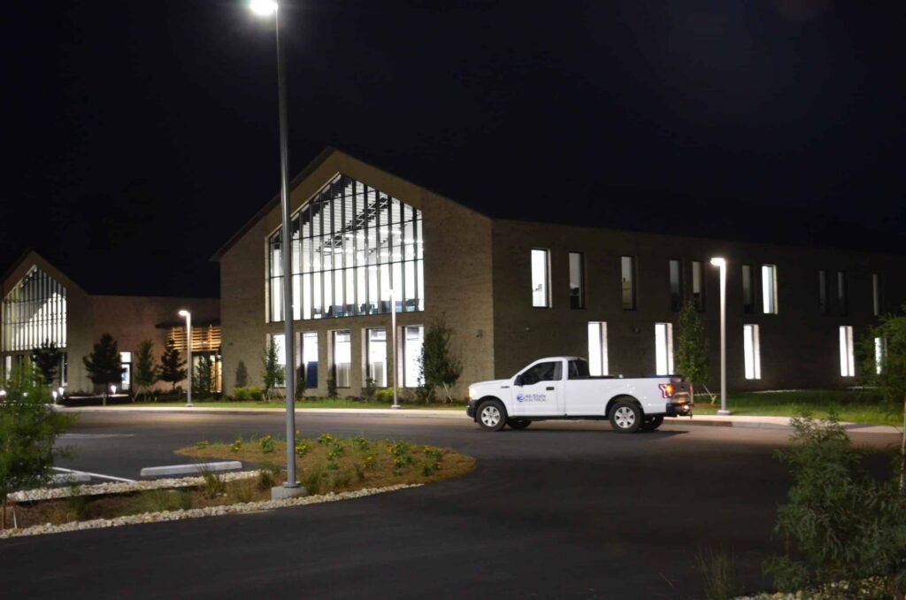 A white pickup truck is parked outside a modern building with large glass windows at night, illuminated by streetlights and interior lighting.