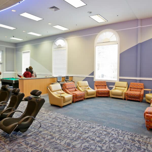 Spacious lounge with patterned and leather chairs in a circle, gaming chairs in front, and people playing pool by large windows.