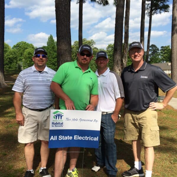 Four men stand outdoors on a golf course, posing behind a sign that reads Habitat for Humanity—All State Electrical. Trees and clear sky are visible in the background.