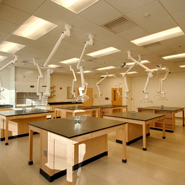 A spacious science lab with wooden benches, black countertops, white ceiling vents, and large windows filling the empty room with light.
