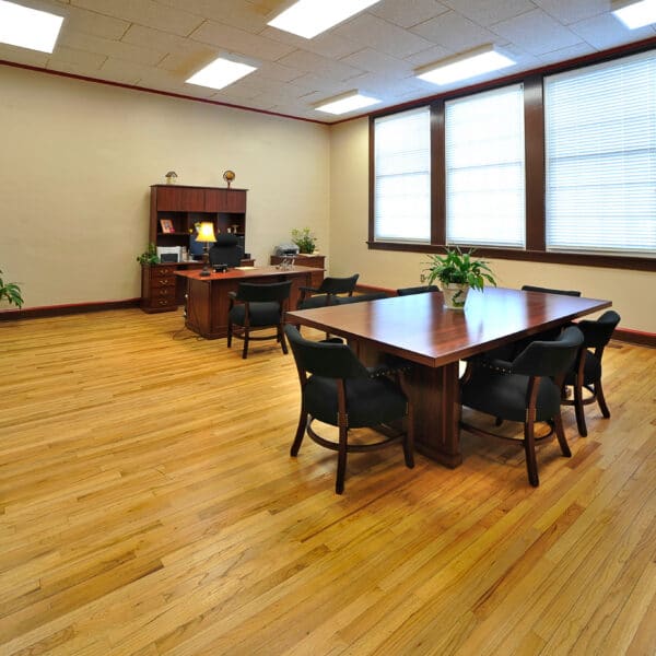 Spacious office with wood floor, big windows, a table with chairs, bookshelves left, back desk with computer, and potted plants.