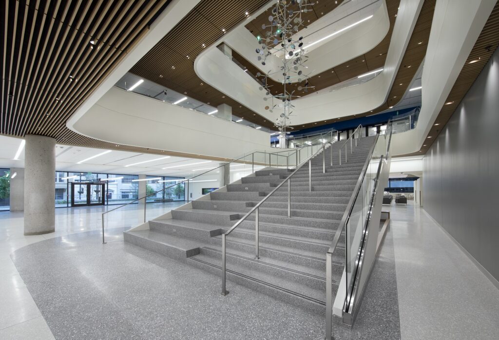 Modern building interior with wide gray stairs, metal railings, terrazzo floors, wooden ceiling panels, large windows, and a hanging abstract sculpture in the atrium. The space is open, bright, and contemporary.