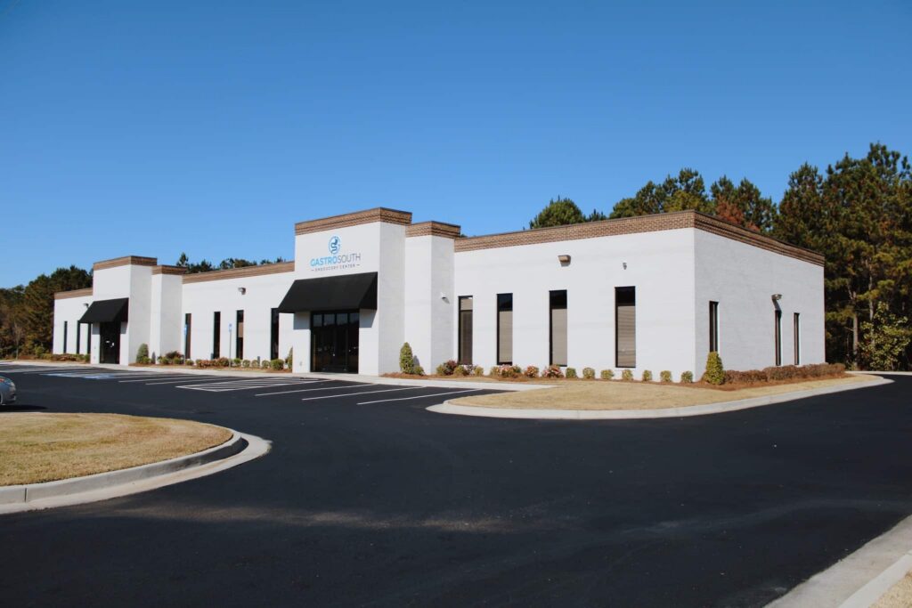 A single-story, white medical office building with large windows and a sign reading Gastro South, surrounded by an empty parking lot and trees under a clear blue sky.