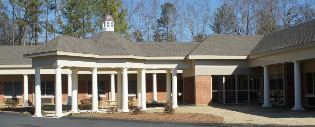 Single-story brick building featuring a columned entrance, peaked roof, and cupola, set among trees with a small seating area in front on a sunny day.