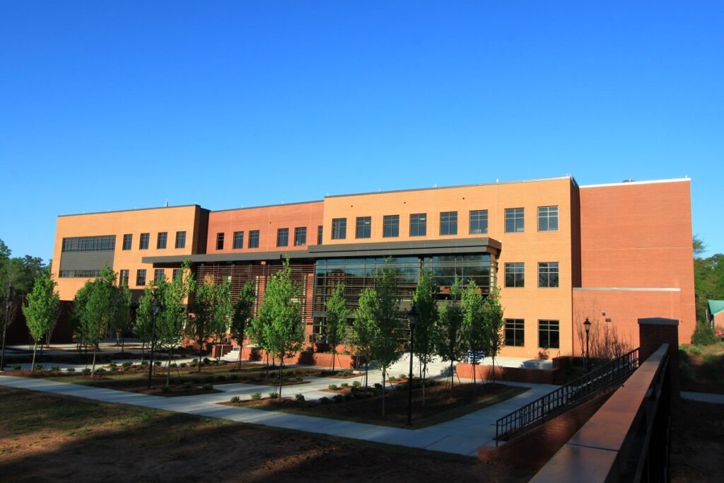 A contemporary three-story brick building featuring expansive windows, bordered by young trees and neatly designed walkways, all beneath a clear blue sky.