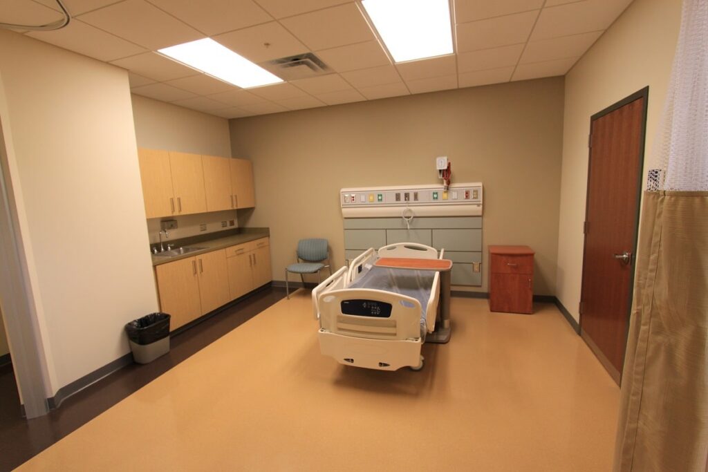 A hospital room featuring a single empty hospital bed, wooden cabinets, a chair, a trash can, and medical equipment mounted on the wall. The room appears clean and brightly lit by overhead lights.