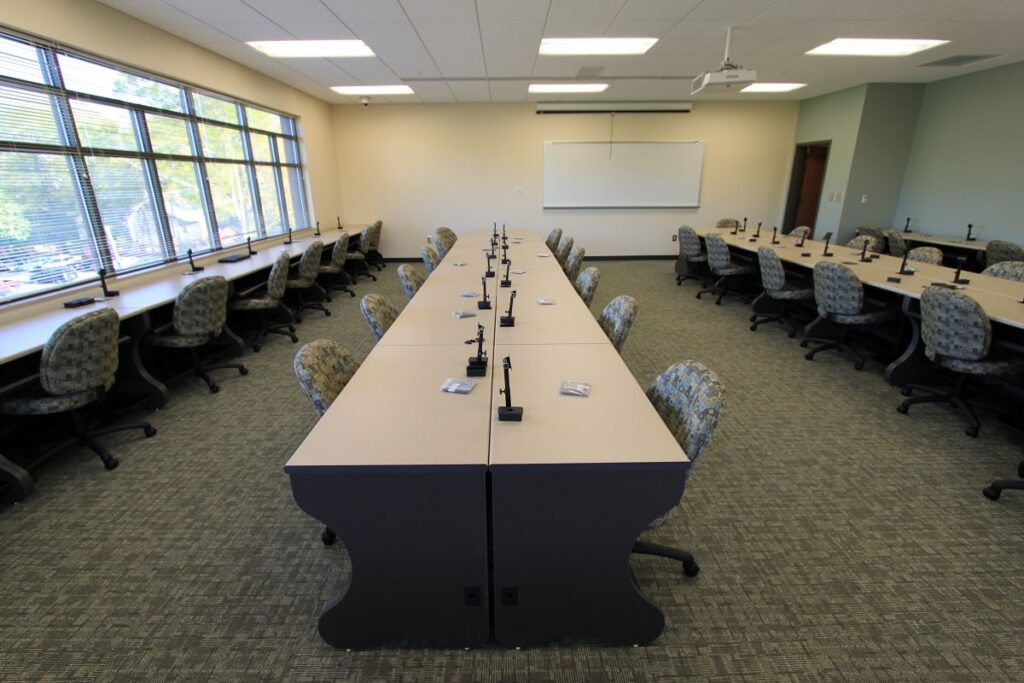 A contemporary classroom features multiple rows of desks paired with office chairs and microphones. Large windows fill the space with natural light, while a whiteboard and projector are mounted at the front of the room.