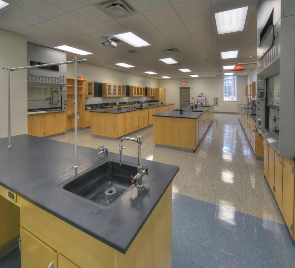 A large, empty science laboratory featuring black countertops, several sinks, wooden cabinetry, and bright overhead lights. Safety equipment and workstations are neatly organized around the space.